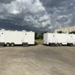 image displays two large, white portable restroom trailers, likely used for events or temporary sites